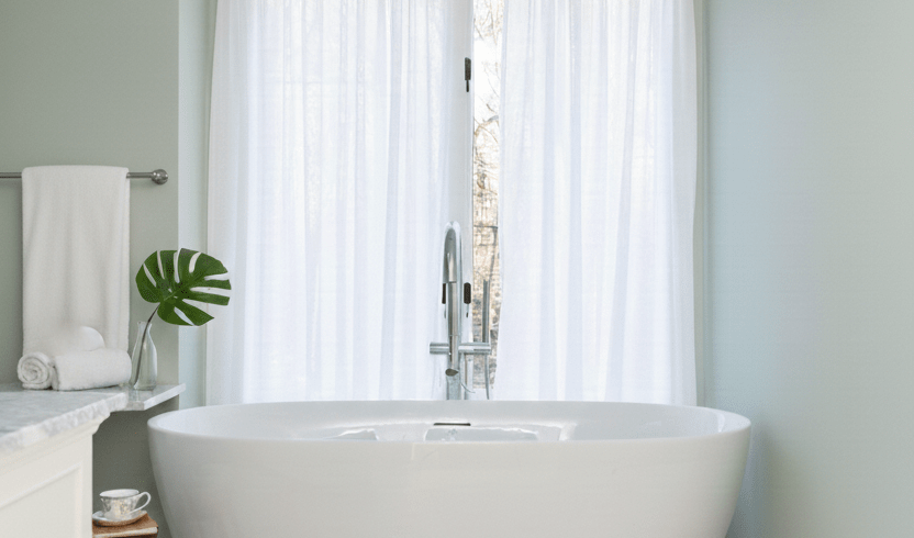 Spa-like master bathroom design in New Haven County with a freestanding soaking tub, sheer white curtains, and marble tile flooring.